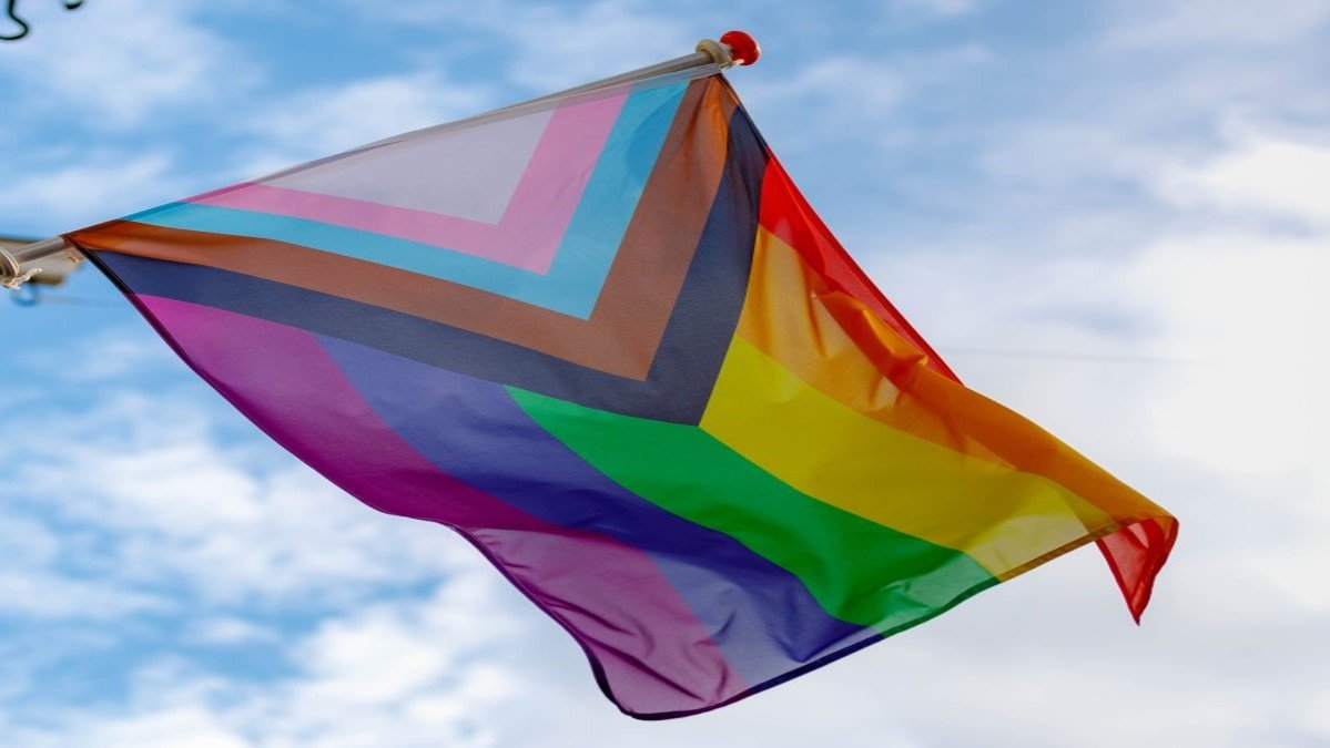 Photograph of a pride flag flying against a blue sky.