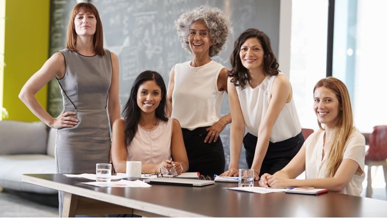 Five women around a table where various writing pads and drinks of water are arranged, all facing the camera.