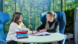 Two female Education students sitting at desk looking at each other with books on table in front