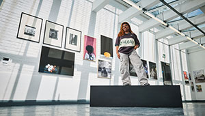 A photograph of Photography graduate Chandni Raithatha standing on top of a black plinth. She is pictured in front of her artwork - a series of framed photographic images a mixture of portraits and images of everyday objects