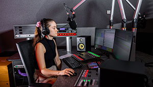 Female student sat operating the control desk in the radio studio. She is wearing headphones focussing on the audio levels