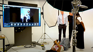 Photoshoot for Prospect Magazine front cover image. A photography student in lying down on her back, in the large white scope in the main photo-studio, and pointing her camera upwards at three people, two male, one female all of them are dressed in business attire. You can also see tripods and spot lights in the image