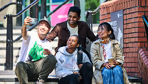 4 students sat on the stairs outside the entrance of the university. It is sunny and they are taking a group selfie
