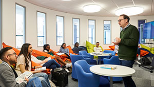 A tutor addressing a group of students seated in a circle around the edge of a classroom.